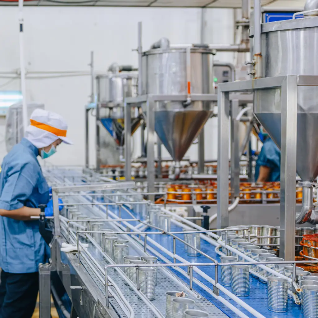 Food manufacturing with large stainless vats and technician at the side with blue top and mask.